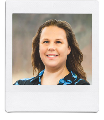 A woman with long brown hair smiles at the camera. She is wearing a dark blue top with a floral pattern. The photo has a white Polaroid-style frame. The background is indistinct and appears blurred.