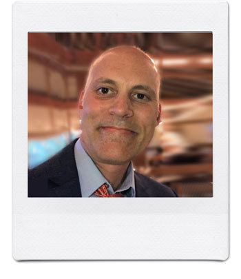 A bald man in a suit and tie is smiling at the camera. The background is blurred with warm, earthy tones, giving the image a cozy ambiance. The photo has a Polaroid-style white border.