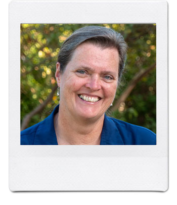 A smiling person with short hair, wearing a dark blue shirt, is pictured outdoors in a setting with greenery in the background. The photo has a white border, resembling a Polaroid picture.