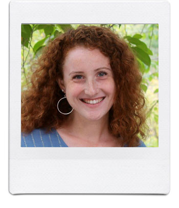 A woman with curly red hair, wearing a blue top and a large circular earring, smiles warmly at the camera. She stands in front of a backdrop of lush, green foliage. The image is framed to resemble a Polaroid photograph.