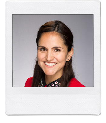 A person with long brown hair and dark eyes smiles at the camera. They are wearing a red top with a floral collar. The background is a neutral gray. The photo is framed to resemble a polaroid picture.
