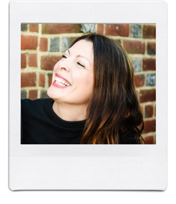 A woman with long dark hair and bright red lipstick is smiling and looking off to the side. She is standing in front of a brick wall and wearing a black top. The photo has a white border, resembling a Polaroid picture.