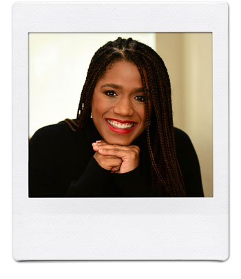 A person with long braided hair, wearing a dark top, is smiling and looking at the camera with their chin resting on their folded hands. The photo has a white border, resembling a Polaroid picture.