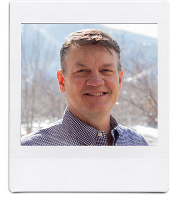 A person with short hair and wearing a checkered shirt smiles while standing outdoors. The background shows snowy trees and a mountainous landscape under a clear sky. The image is framed like a Polaroid photo.