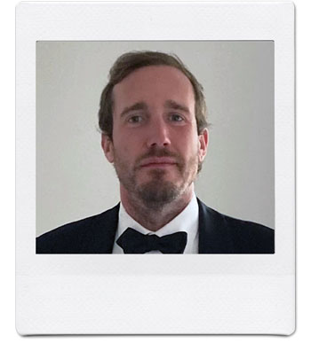 A man with short brown hair and a beard is dressed in a black tuxedo with a black bow tie. He is looking directly at the camera against a plain, light-colored background, framed in a white Polaroid-style border.