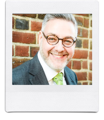 A smiling man with gray hair and a beard, wearing glasses, a suit, and a green tie, stands against a brick wall. The photo has a Polaroid-style border.