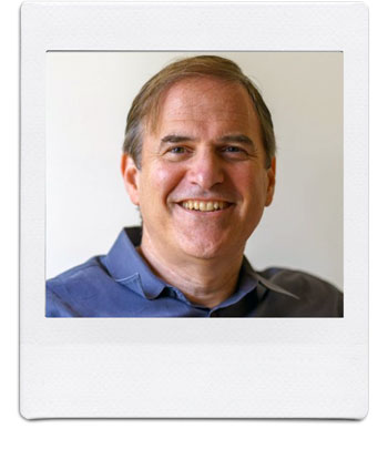 A portrait of a middle-aged man with short brown hair, smiling at the camera. He is wearing a blue collared shirt. The photograph has a white Polaroid-style frame. The background is plain and out of focus.