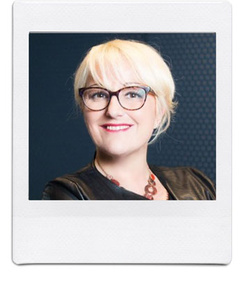 A person with short, blonde hair is smiling and wearing glasses. They are dressed in a dark outfit and have a necklace with circular beads. The photo has a Polaroid-style border. The background is dark with a subtle textured pattern.