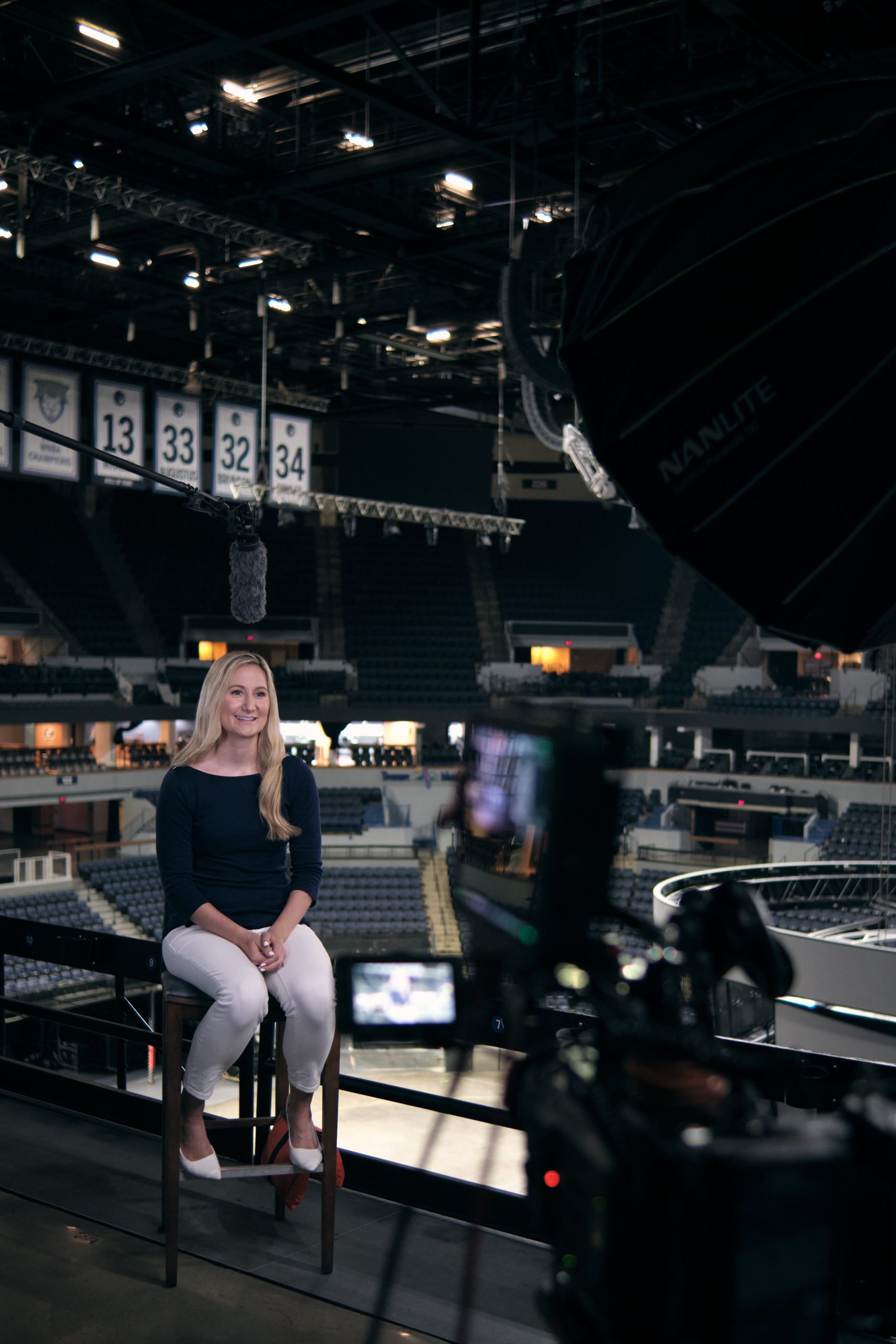 A woman with long blonde hair sits on a stool in an arena, being filmed for an interview. Stage lights, camera equipment, and stadium banners are visible in the background.