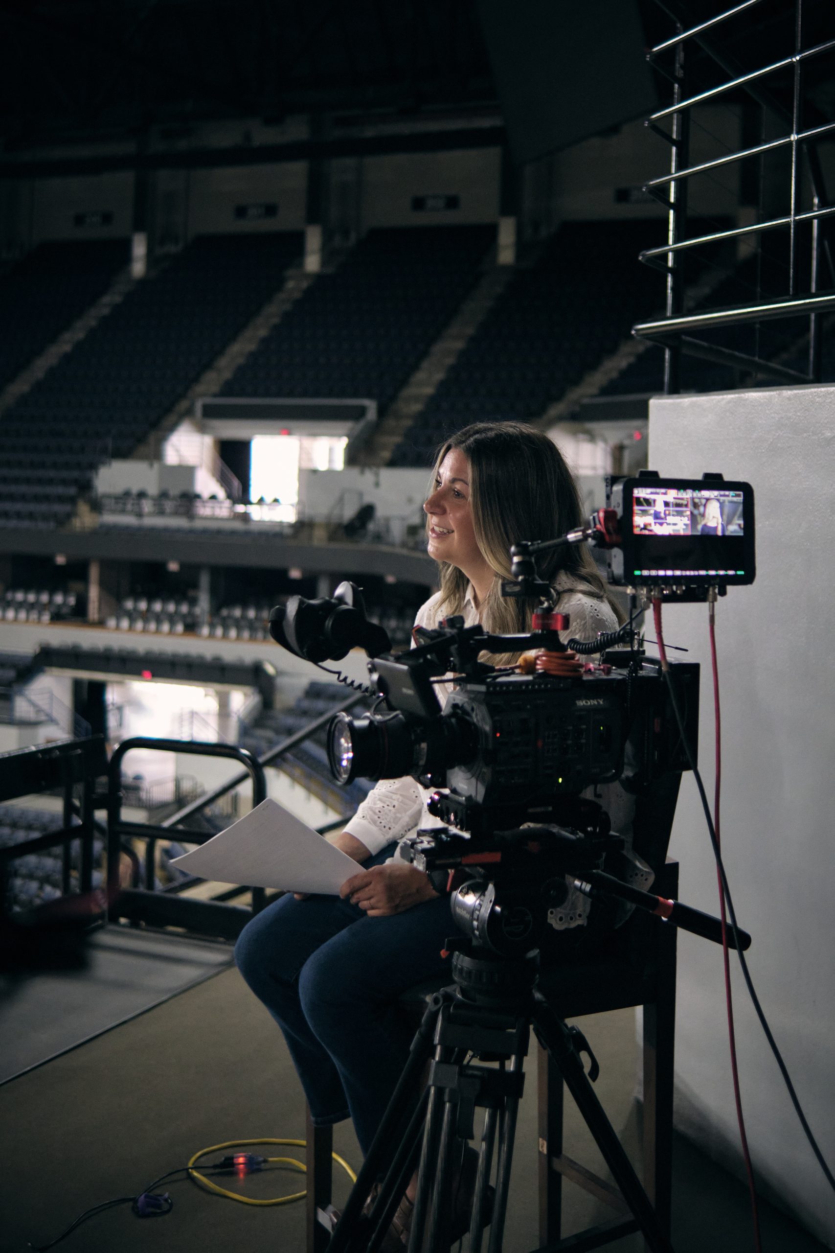 A woman sits in an empty stadium, smiling beside a professional video camera on a tripod, with a monitor attached, holding papers as she prepares for an interview or recording.