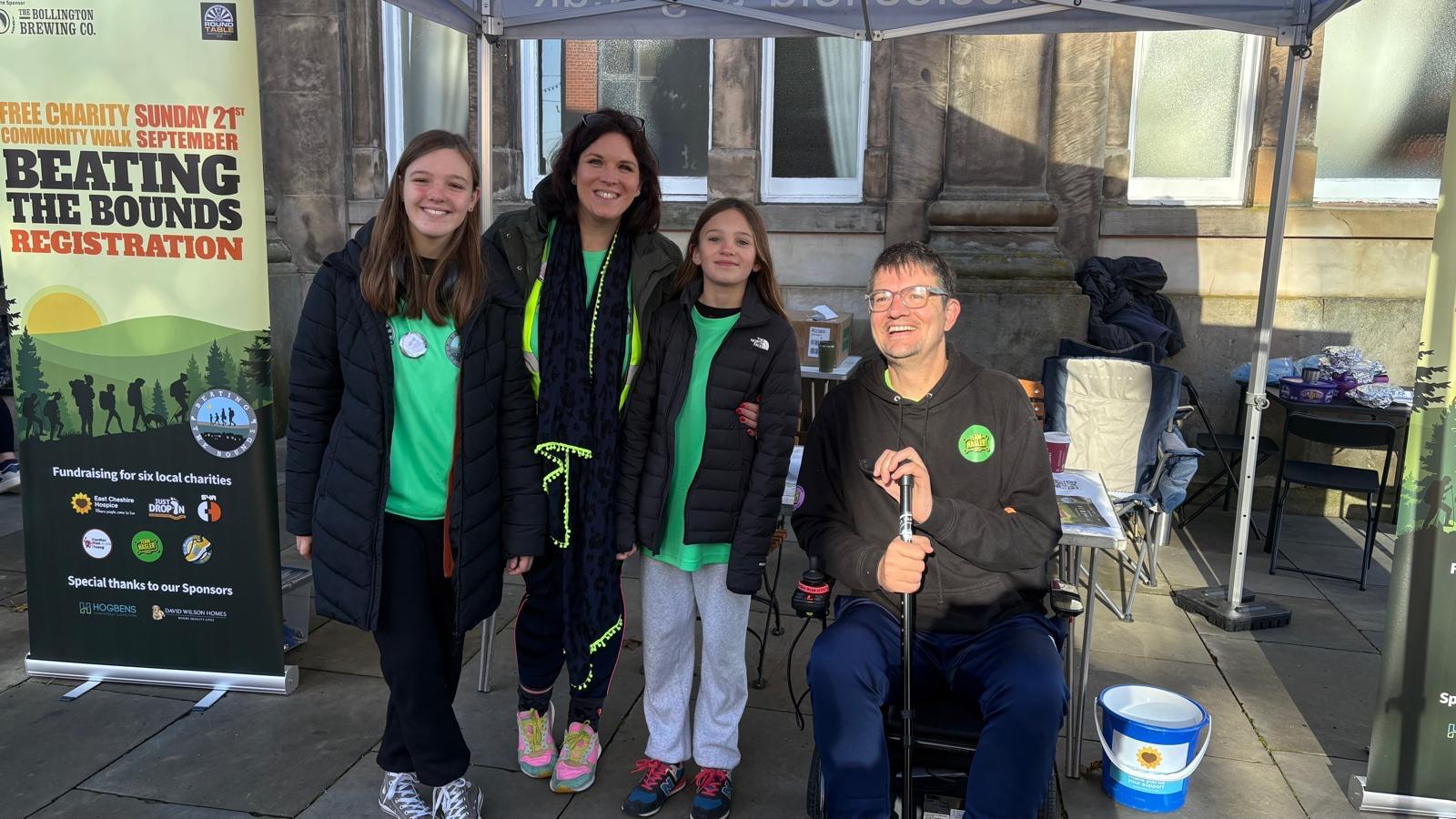 Four people, three standing and one seated in a mobility scooter, smile under a tent at an outdoor event. Banners behind them promote "Beating the Bounds Registration"—a charity event inspiring all to live your life fully.