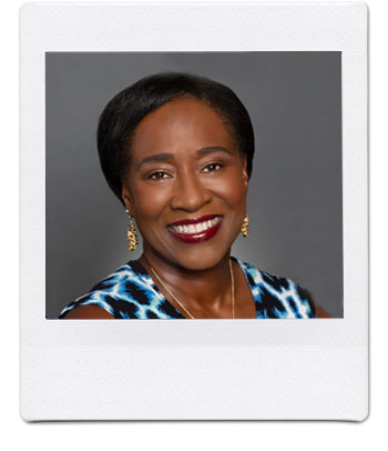 A woman with short black hair smiles warmly in a portrait. She is wearing a sleeveless blue, black, and white patterned top and gold earrings. The background is a plain gray. The photo has a white, Polaroid-style border.