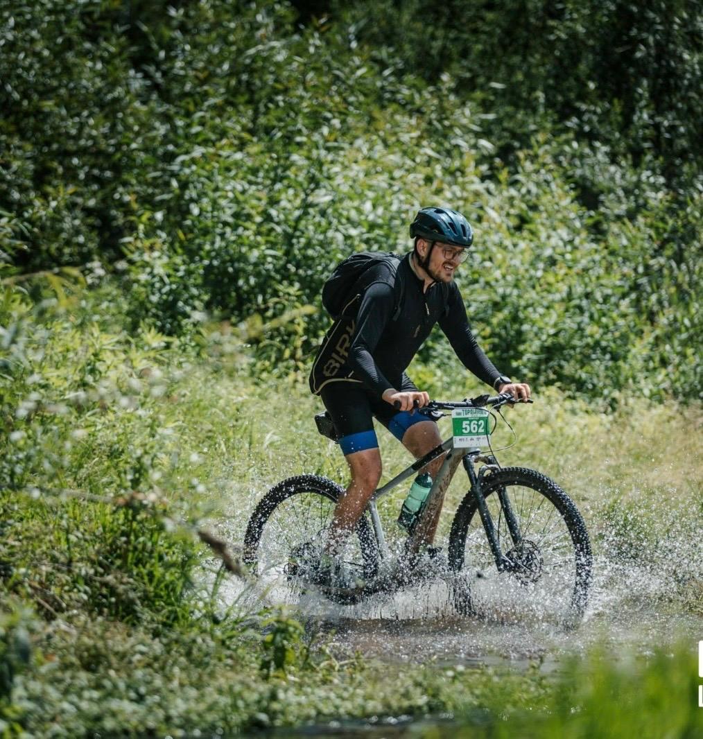 A cyclist wearing a helmet, sunglasses, and a backpack rides a mountain bike through a shallow, splashing stream surrounded by lush green vegetation. The bike displays a number plate reading "562.