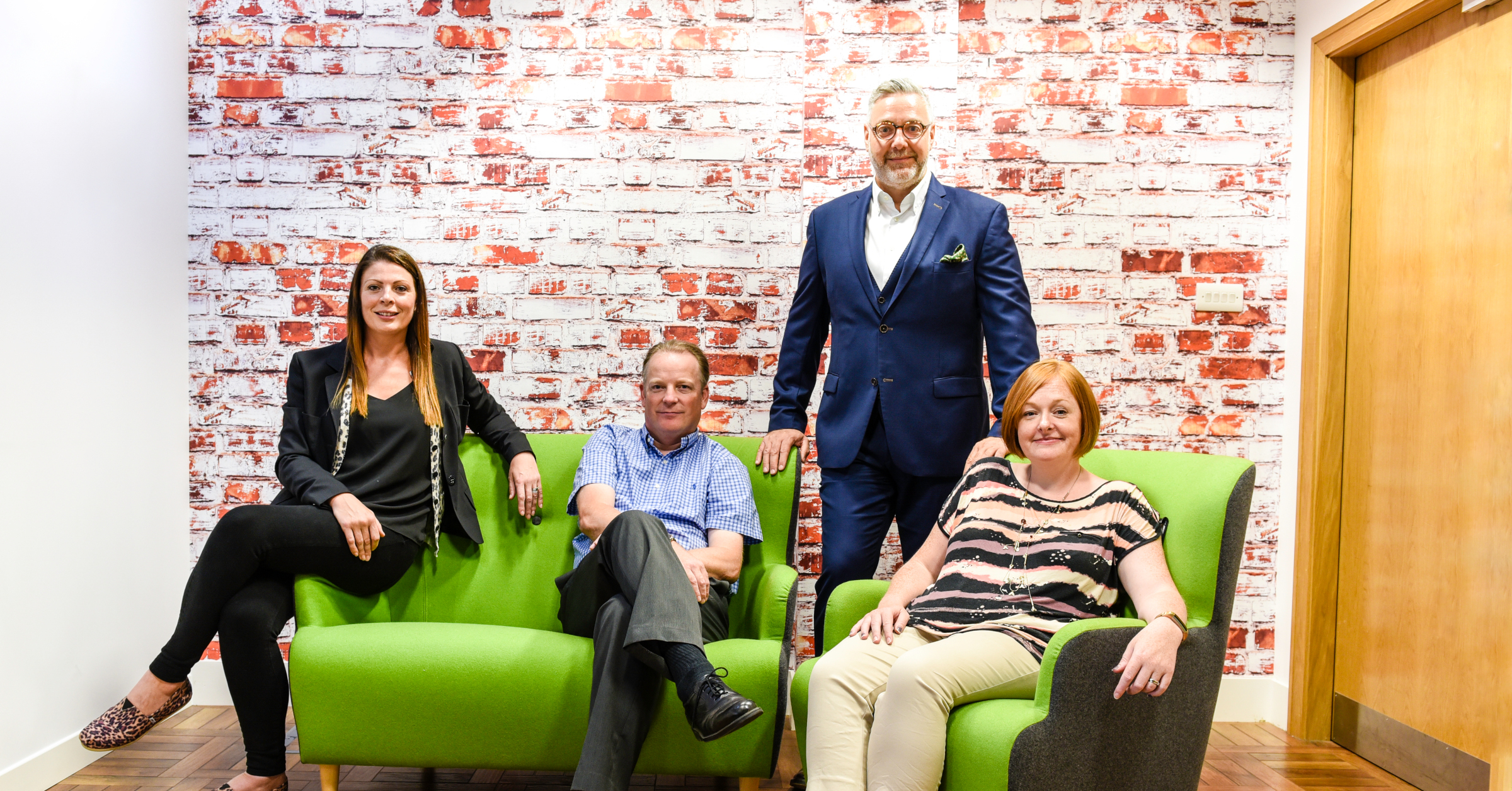 Four people are posing in an office setting with a brick wall backdrop. Two are seated on green chairs, and two are standing behind. They are dressed in business-casual attire, with one person wearing a suit.