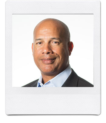 A man with a shaved head and a neutral expression is wearing a dark suit jacket and a light blue dress shirt. He is looking directly at the camera against a plain white background. The image is framed like a Polaroid photo.
