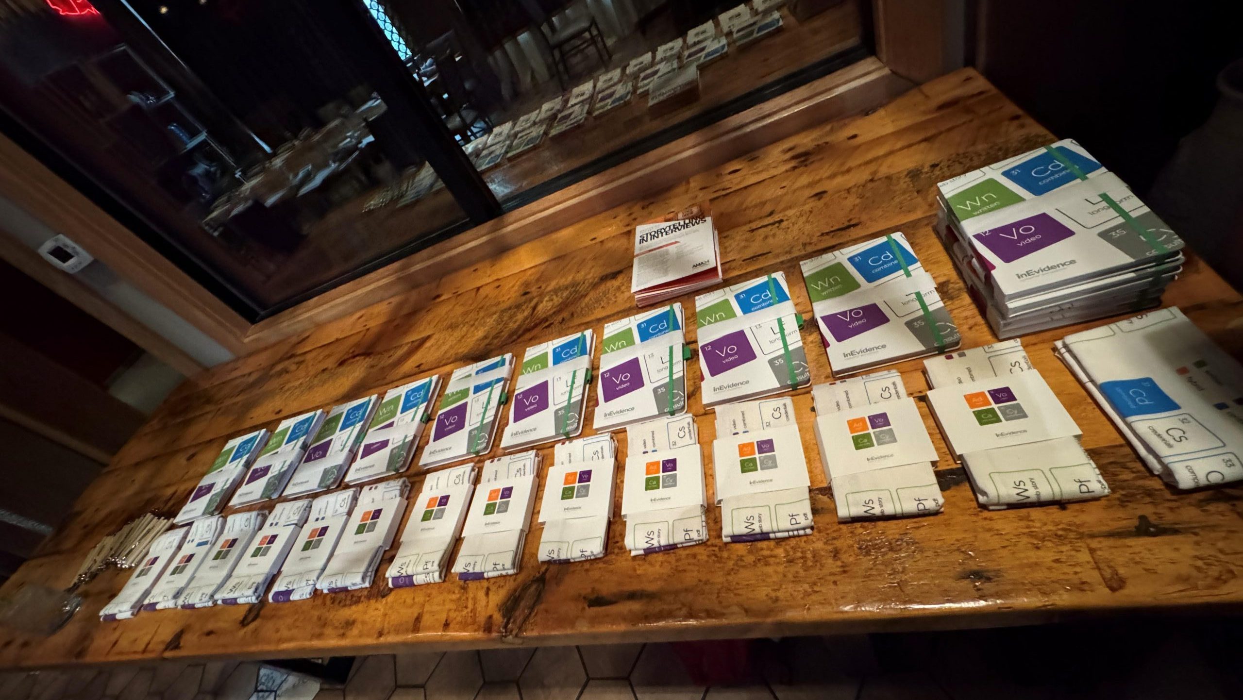 A wooden table is covered with neatly arranged stacks of cards. Each card features colorful labels and logos, likely indicating different categories or types. The background shows a tiled floor and a window with reflections of a dimly lit interior space, reminiscent of the ambiance at AMAx Raleigh '24.