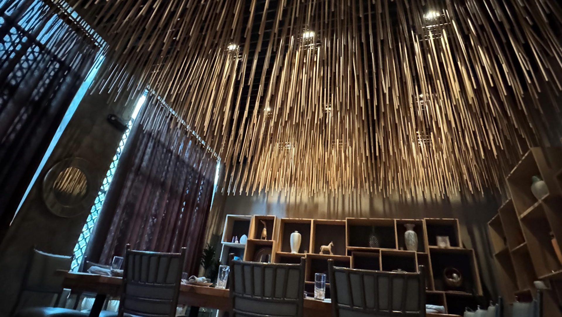 A dining room with a striking ceiling design featuring numerous bamboo poles hanging vertically. The room is dimly lit, with a large wooden dining table surrounded by chairs. Shelving with decor items lines the walls, creating a cozy and intimate atmosphere perfect for planning your AMAx Raleigh 2024 experience.