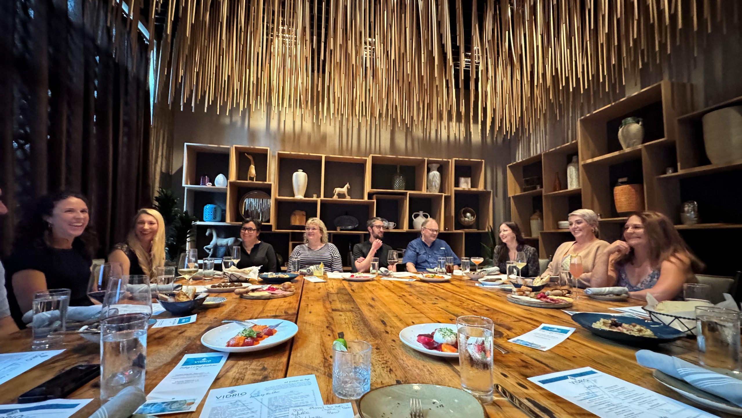 A group of people sits around a large wooden dining table in a stylish restaurant, AMAx Raleigh 2024, with a unique ceiling design featuring numerous hanging wooden rods. Plates of food, drinks, and menus are visible on the table. Shelves with decorative items line the back wall.