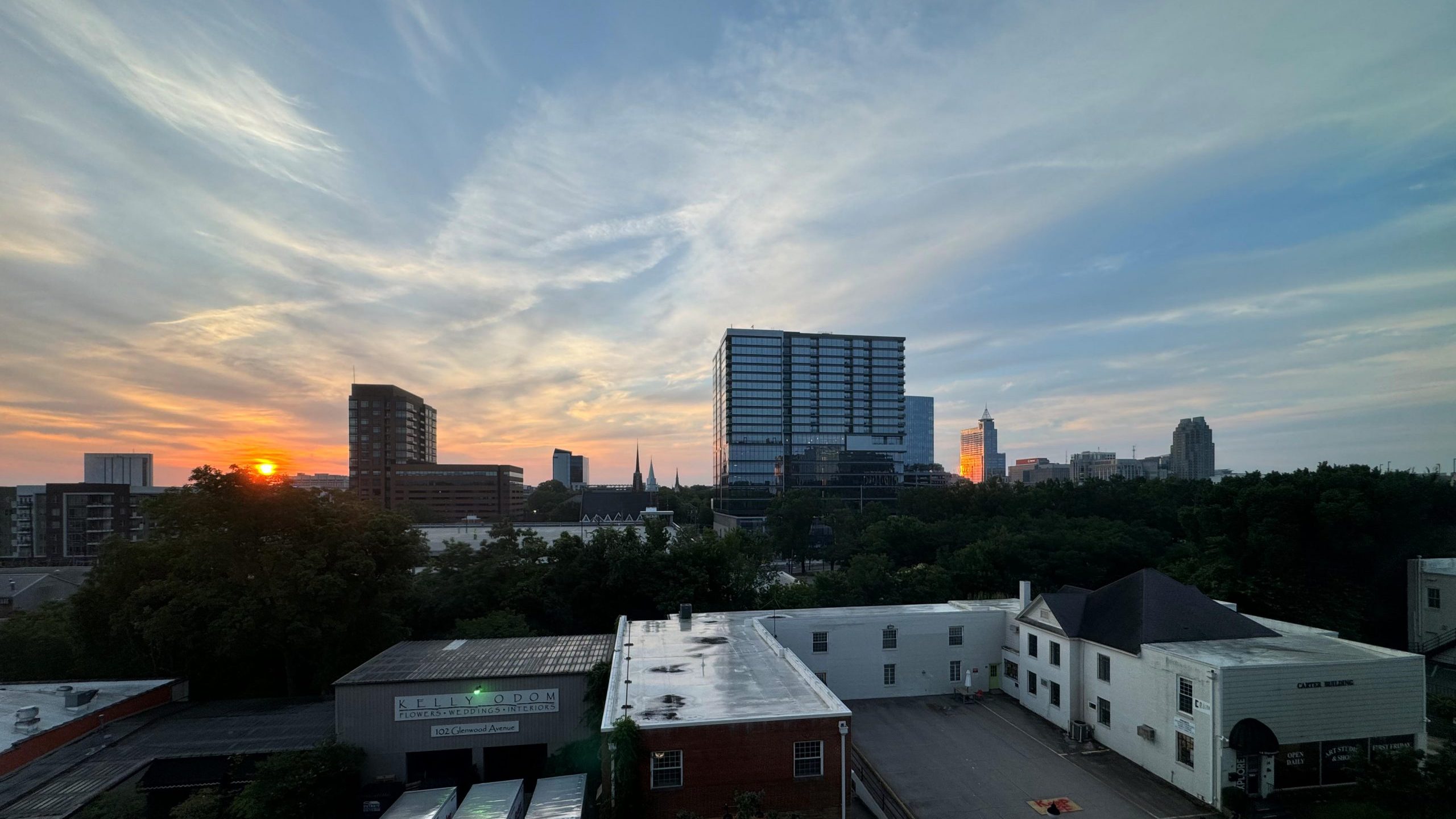A cityscape view at sunset, featuring a mix of buildings and greenery in Raleigh '24. The sun is partially visible on the horizon, casting an orange hue across the sky. Several buildings, including a prominent tall building, and parked white vans are in the foreground.