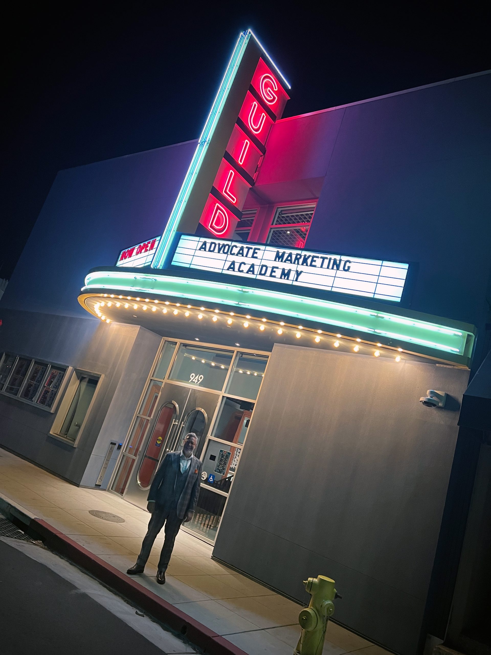 A person stands in front of a building with a neon sign that reads "GUILD." The marquee below displays the message "ADVOCATE MARKETING ACADEMY." The scene is illuminated by the building's lights, and it appears to be nighttime. A fire hydrant is visible on the sidewalk.