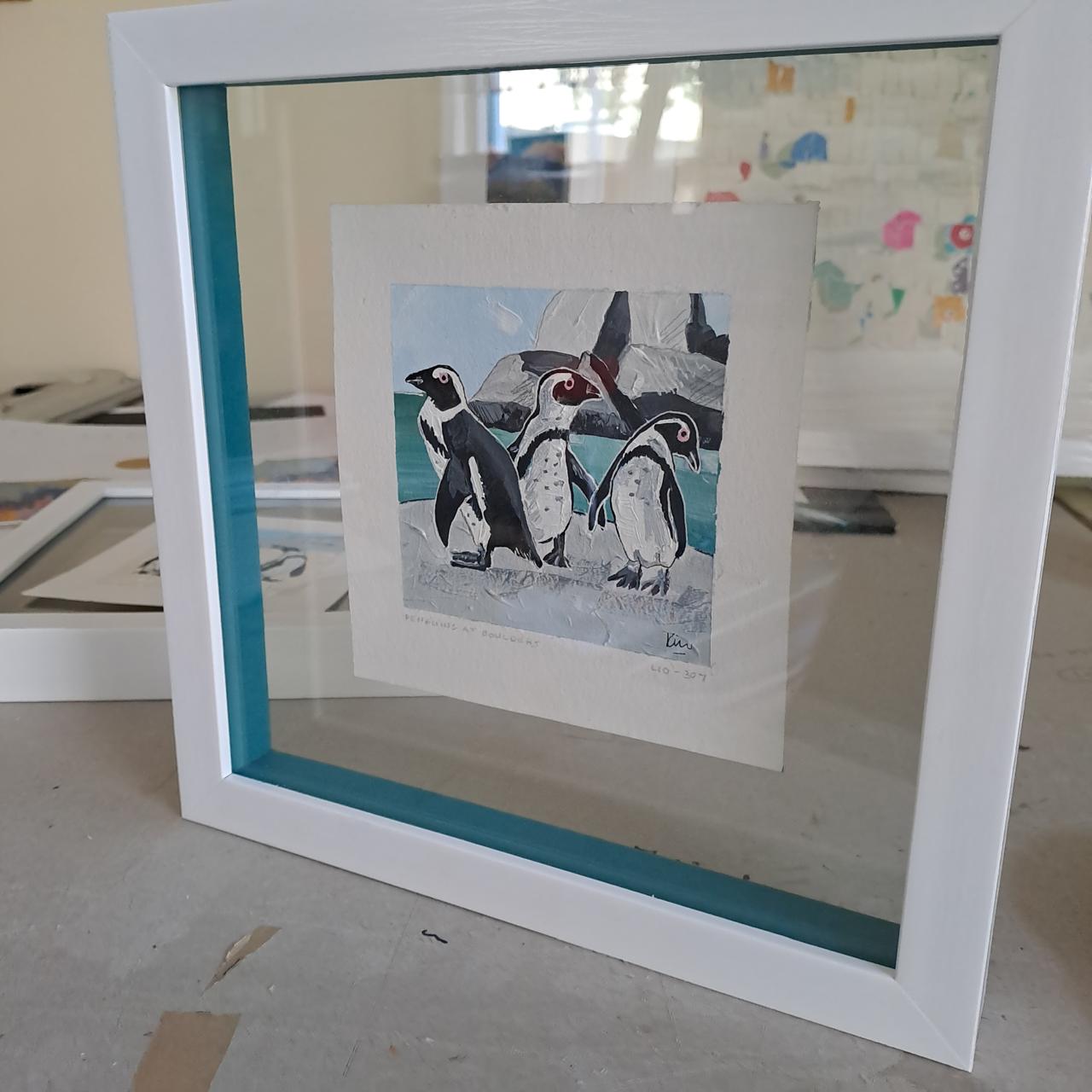 A framed painting of three penguins standing on ice against a backdrop of rocks and water. The artwork is displayed inside a white frame with blue trim, set on a table. Other framed pieces are visible in the background.