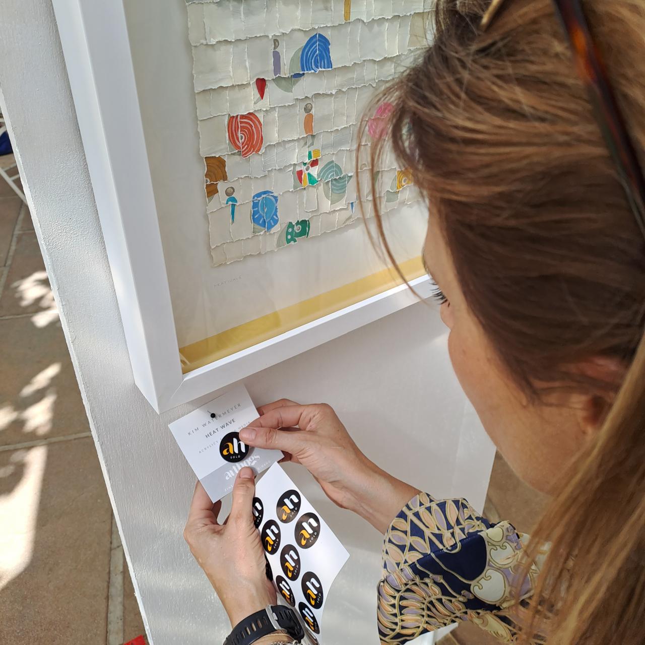 A person places black and gold stickers with "au" on a white surface near a framed artwork with abstract designs. They are wearing a patterned blouse and a black watch.