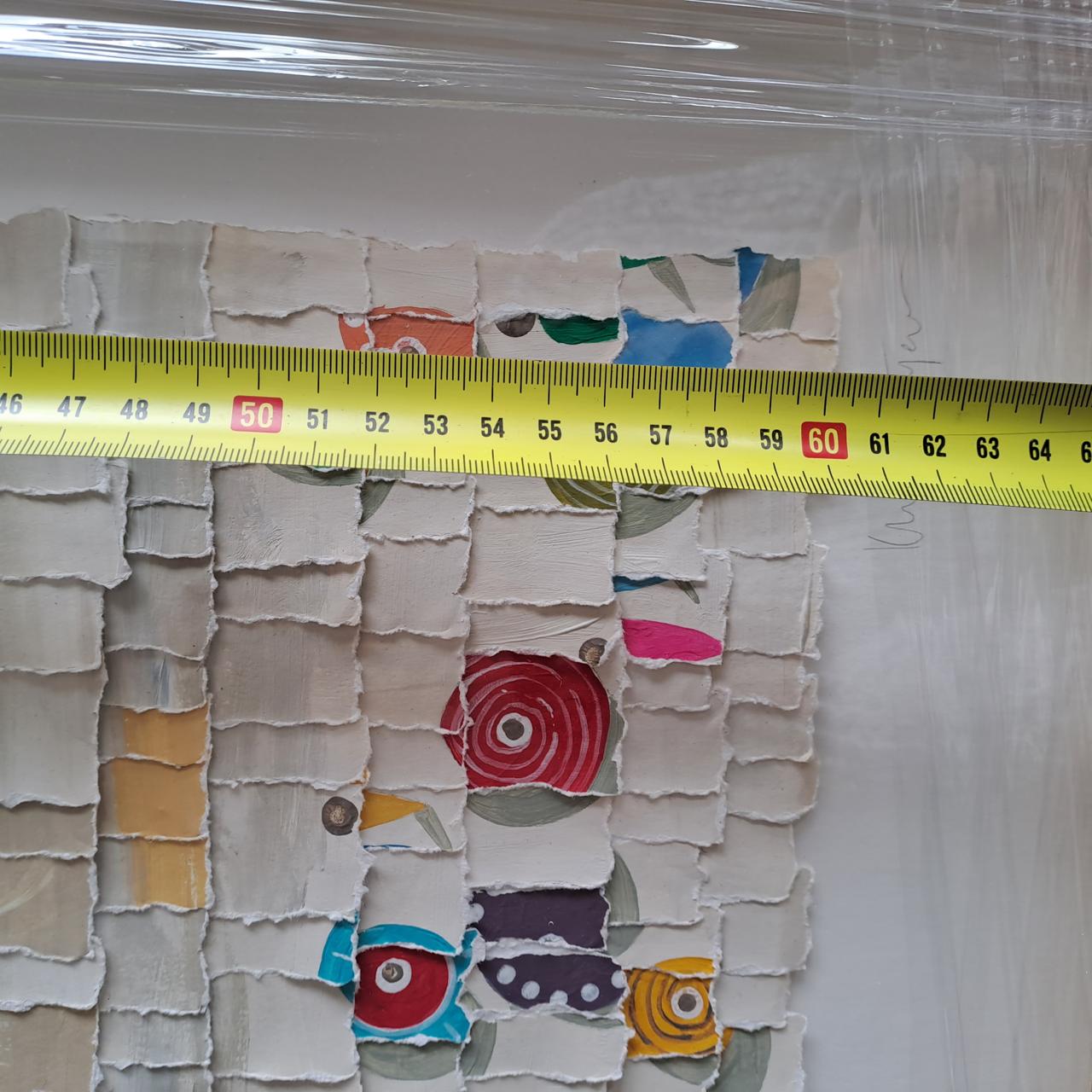 A close-up of a measuring tape over a textured surface with overlapping paper squares in various colors and patterns, including circles and geometric shapes. The tape is stretched across the surface, reading just over 60 centimeters.