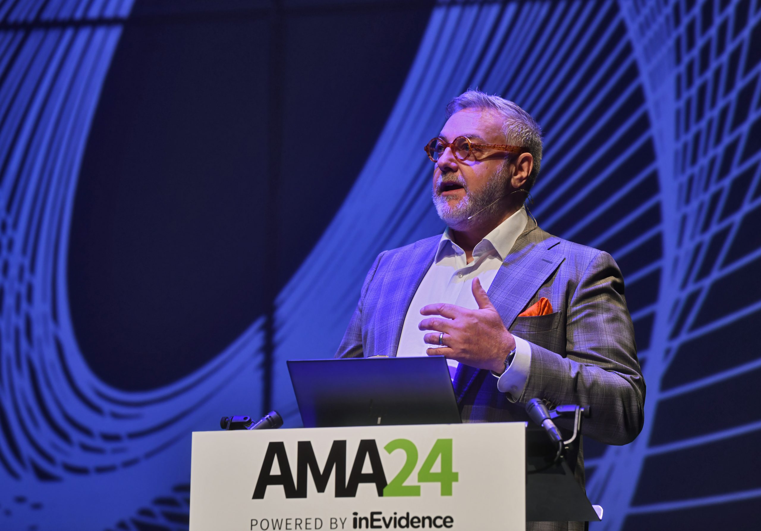 A man with a neatly trimmed beard and glasses stands at a podium during an event. He is wearing a checkered suit with an orange pocket square and is speaking into a microphone. The podium displays a sign with "AMA24" and "Powered by inEvidence" on it. The background features abstract curved patterns.