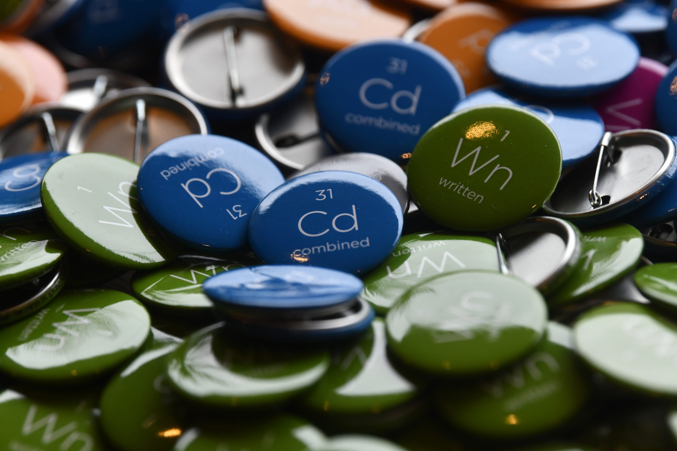 A close-up photograph of various colorful buttons scattered on a surface. The buttons display chemical element symbols and names, such as green ones for "Wn written" and blue ones for "Cd combined." The pile includes buttons in green, blue, and orange.