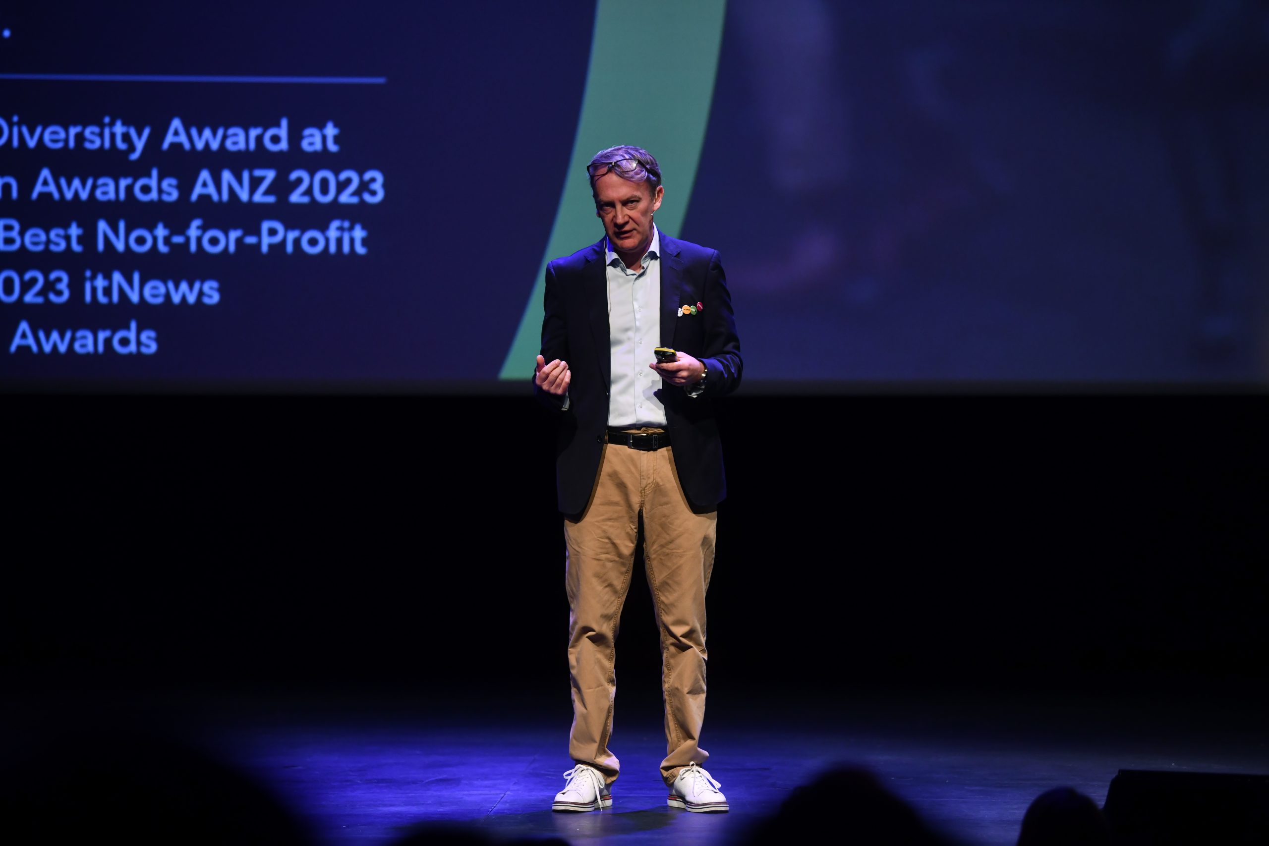 A man in a blazer, khakis, and white sneakers stands on a dimly lit stage, speaking with hand gestures. Behind him, a large screen displays text listing various awards, including the 2023 Diversity Award and Best Not-for-Profit.