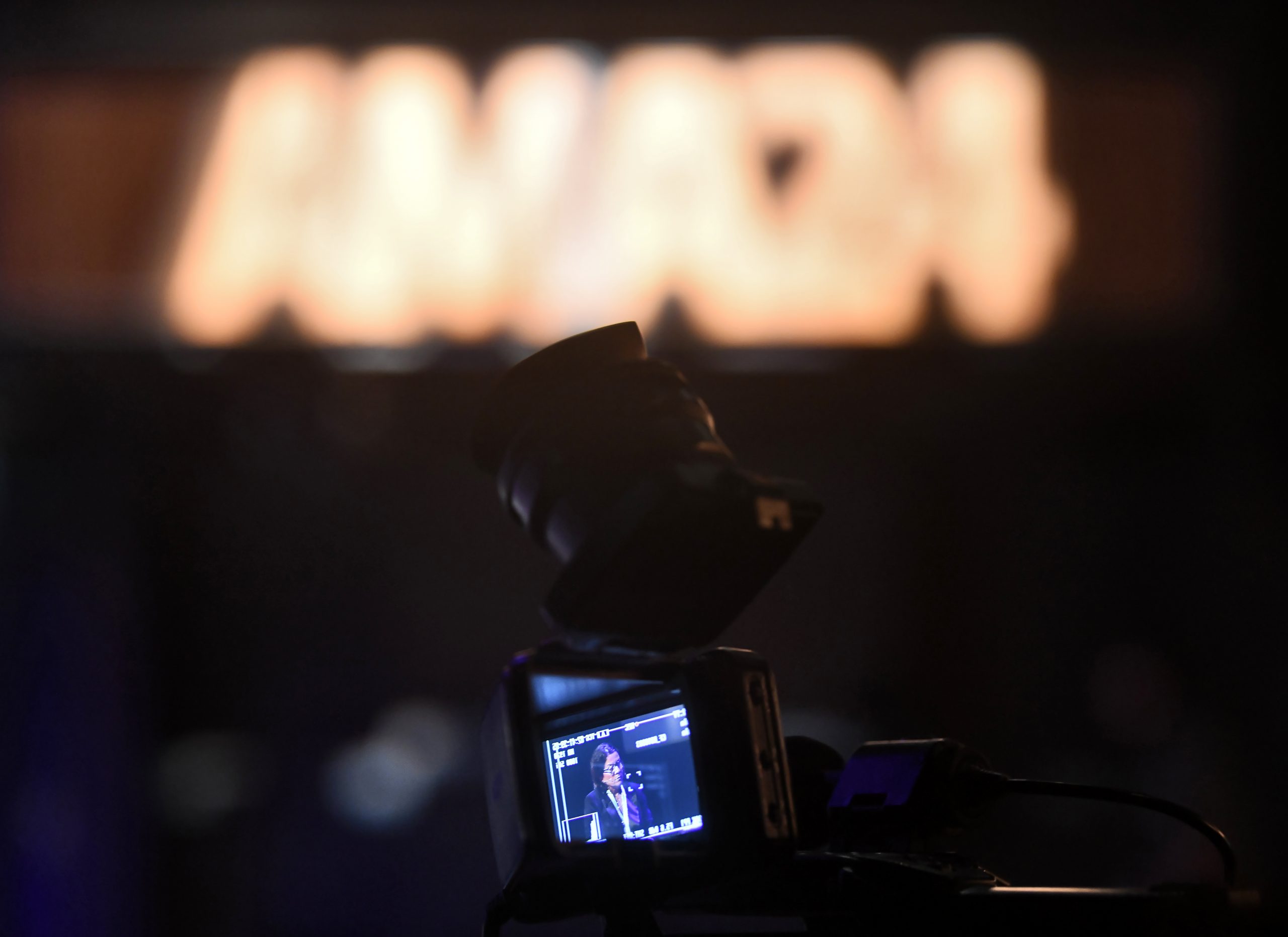 A close-up of a video camera display showing a person speaking on stage. The background is blurred with an illuminated sign that reads "AMADA". The focus is on the camera's screen, capturing the speaker.
