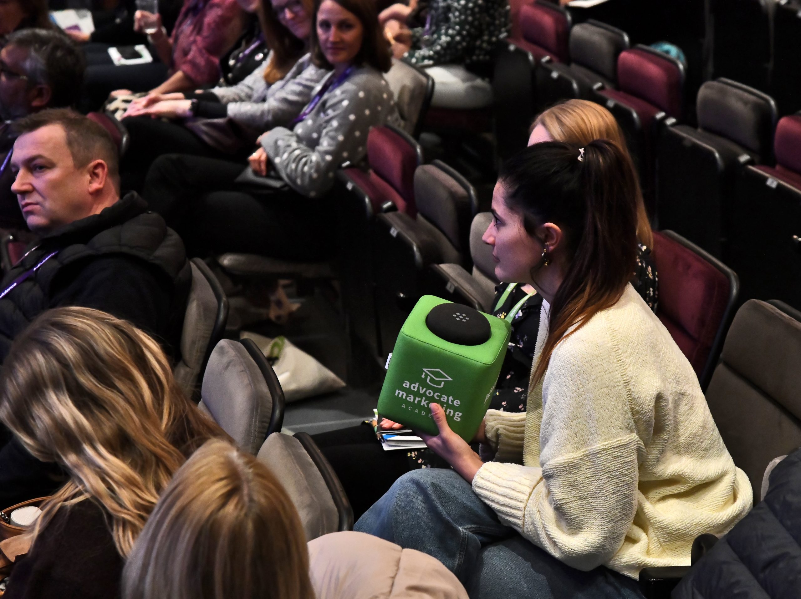 A woman in a white sweater sits among a group of people in an auditorium. She holds a green cube-shaped object with "advocate marketing" written on it. The audience seems attentive, with some looking towards the woman, indicating she might be speaking or asking a question.