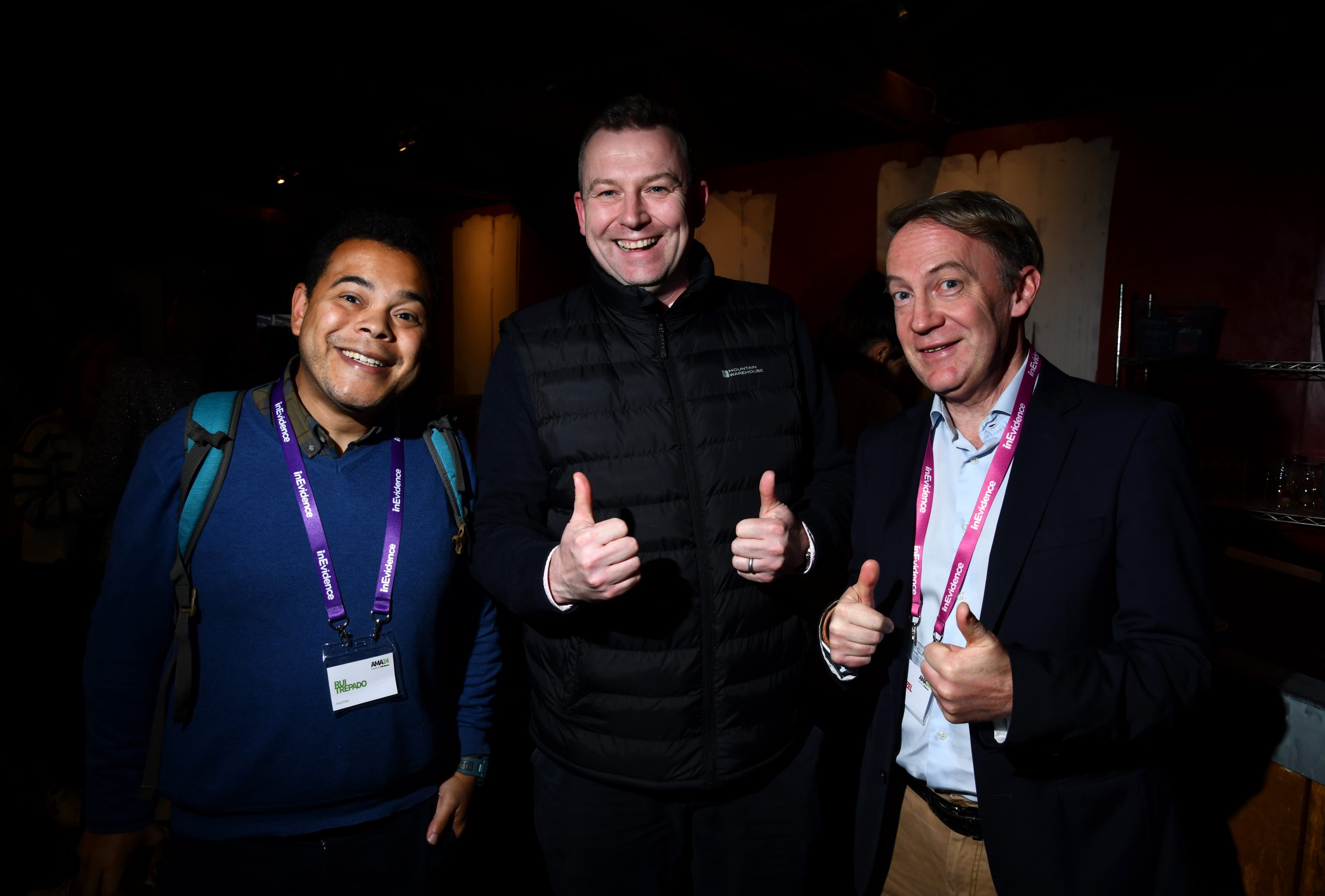 Three smiling men are wearing lanyards and standing closely together in a dimly lit space. The man in the middle and the man on the right are giving a thumbs-up gesture. They appear to be at a social or networking event.