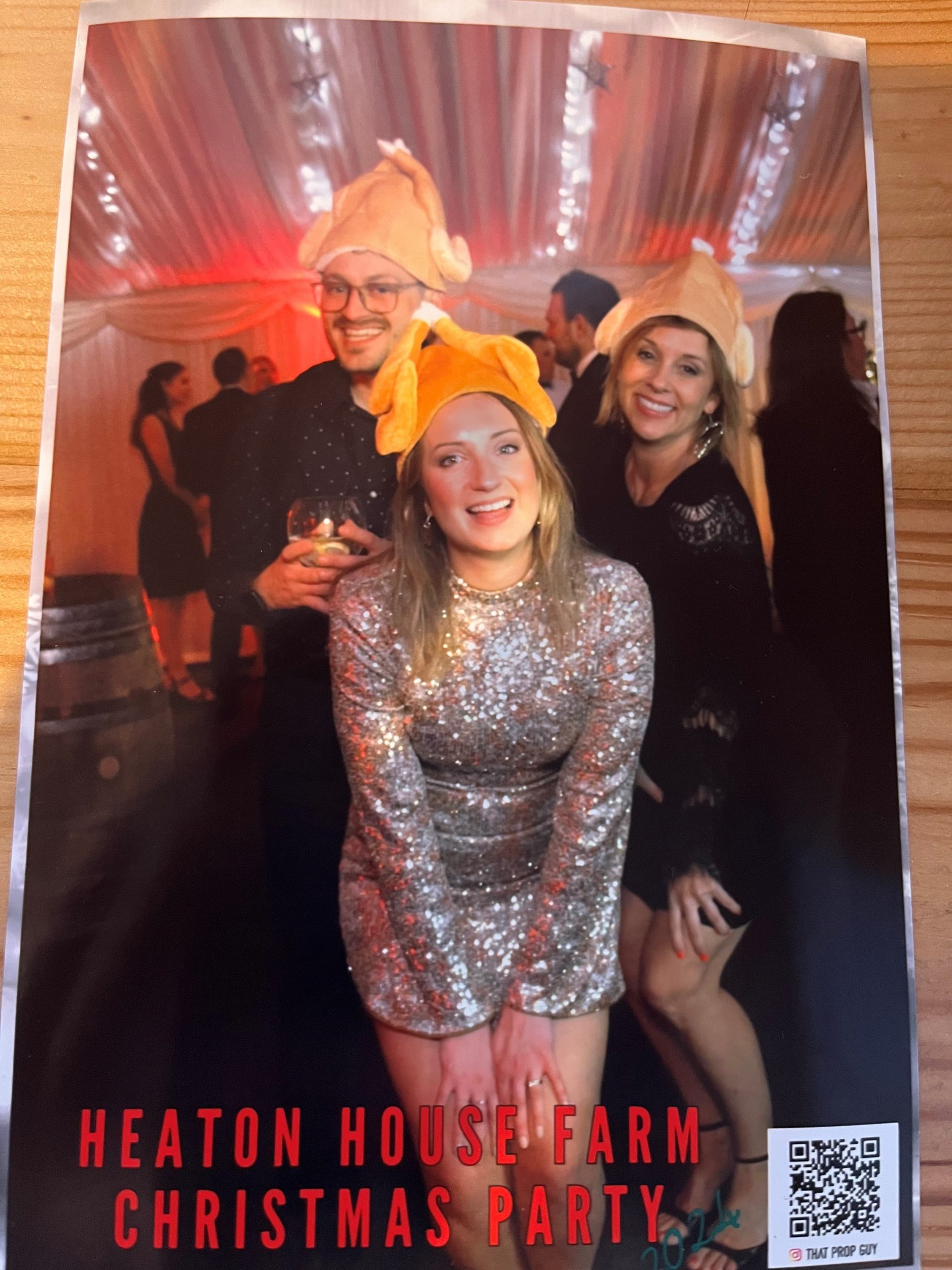 Three people at a party wear turkey-shaped hats and festive outfits, smiling for the camera. "Heaton House Farm Christmas Party" is written at the bottom in red text. Other guests are visible and the venue is decorated with lights.