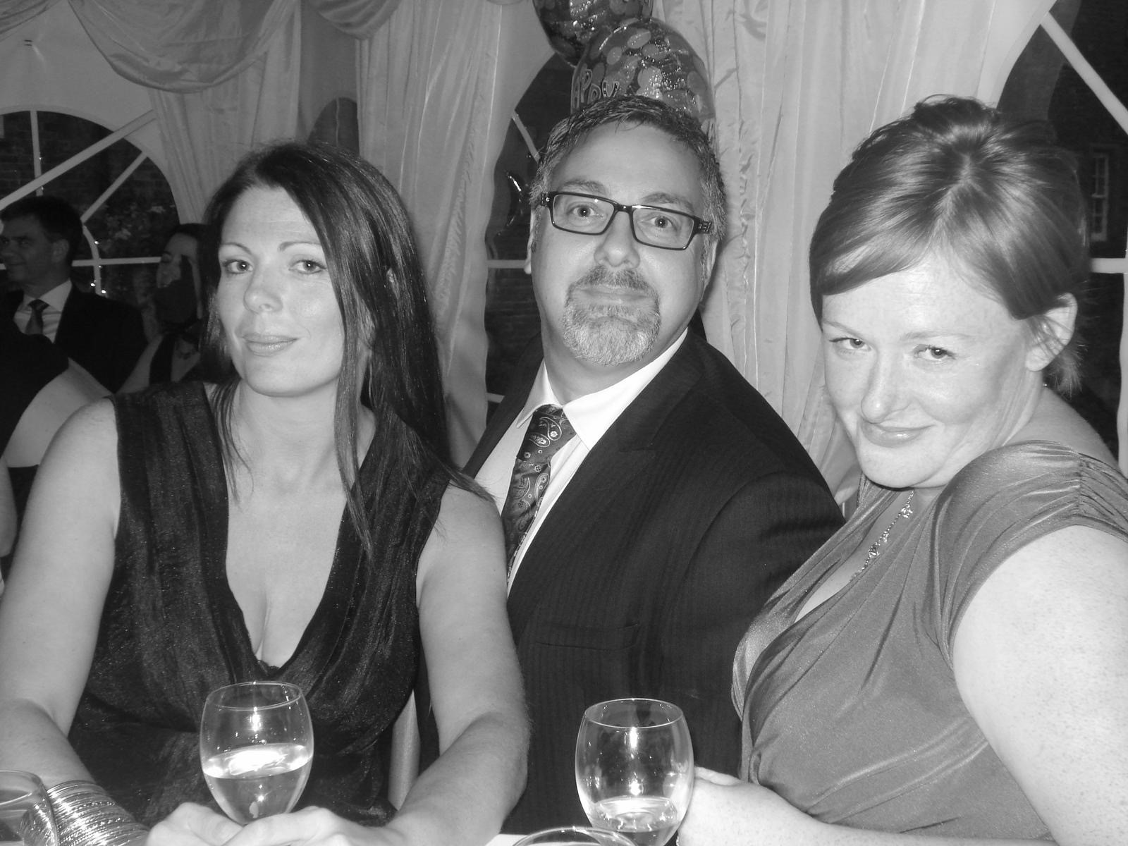 Three people are sitting close together at a formal event, each with a glass of wine. They are dressed in evening attire, with the man in the middle wearing glasses. Draped curtains and a decorative balloon are in the background. The image is in black and white.