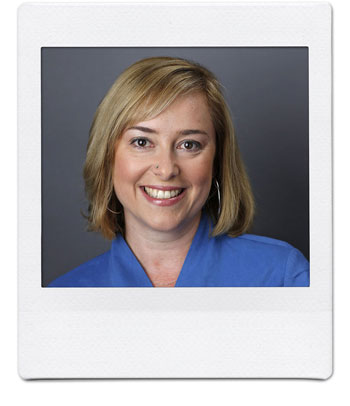A woman with shoulder-length blonde hair is smiling at the camera. She is wearing a blue top and hoop earrings, set against a plain dark background. The photo has a white Polaroid-style border.