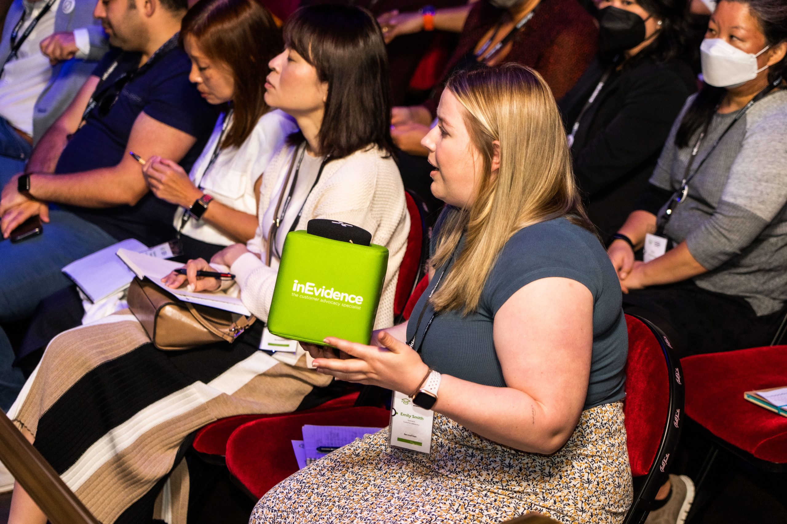 A woman sitting in an audience holds a green microphone cube labeled "InEvidence" as she speaks. Other attendees, seated around her, are listening attentively, taking notes, or wearing masks. They are at an indoor event, likely a seminar or conference.