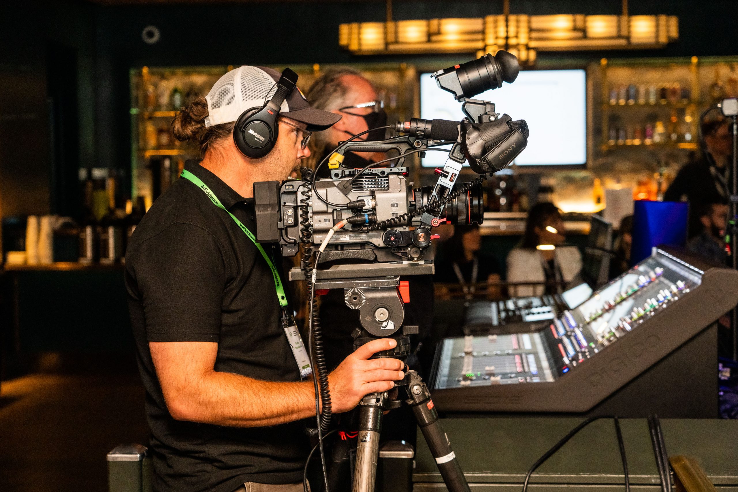 A cameraman wearing a headset and a cap operates a professional video camera mounted on a tripod. Another individual adjusts audio equipment nearby. The background features a dimly lit bar area with shelves of bottles and hanging lights.