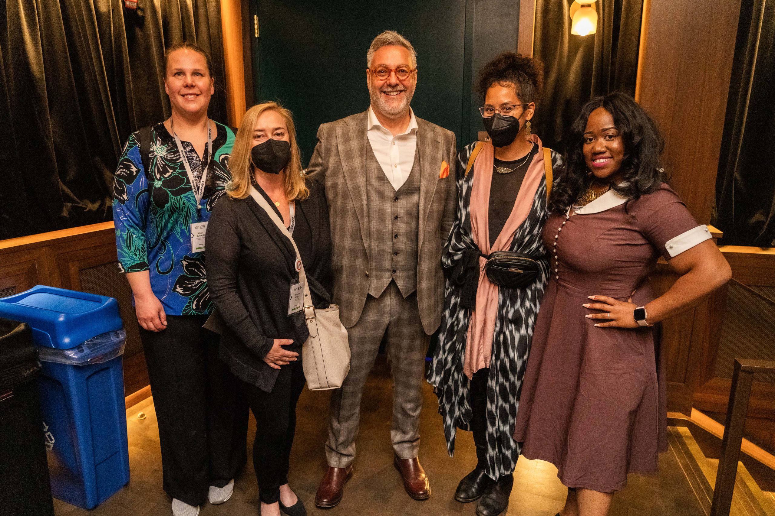 A group of five people standing indoors. The central figure is a man in a checked suit, flanked by four women, some of whom are wearing masks. They all appear to be at an event and are smiling for the camera.