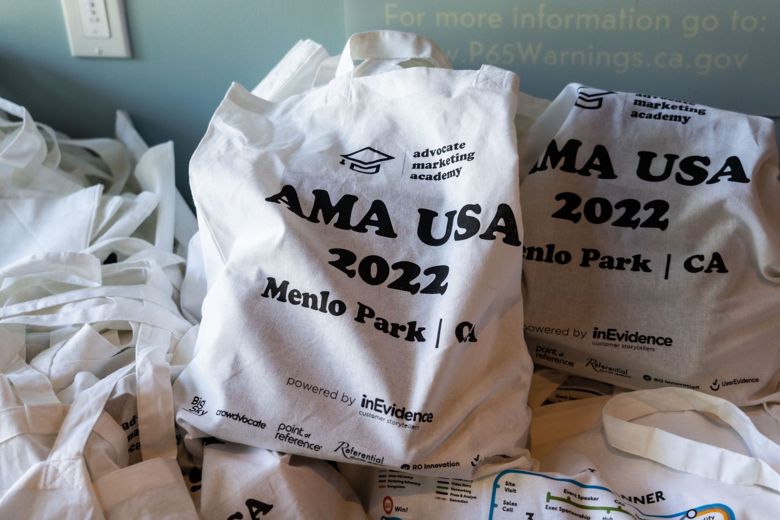 Several white cloth bags printed with "AMA USA 2022 Menlo Park, CA" are piled together. The bags also display various logos and text that mentions "Advocate Marketing Academy" and "powered by InEvidence." More bags can be seen in the background.