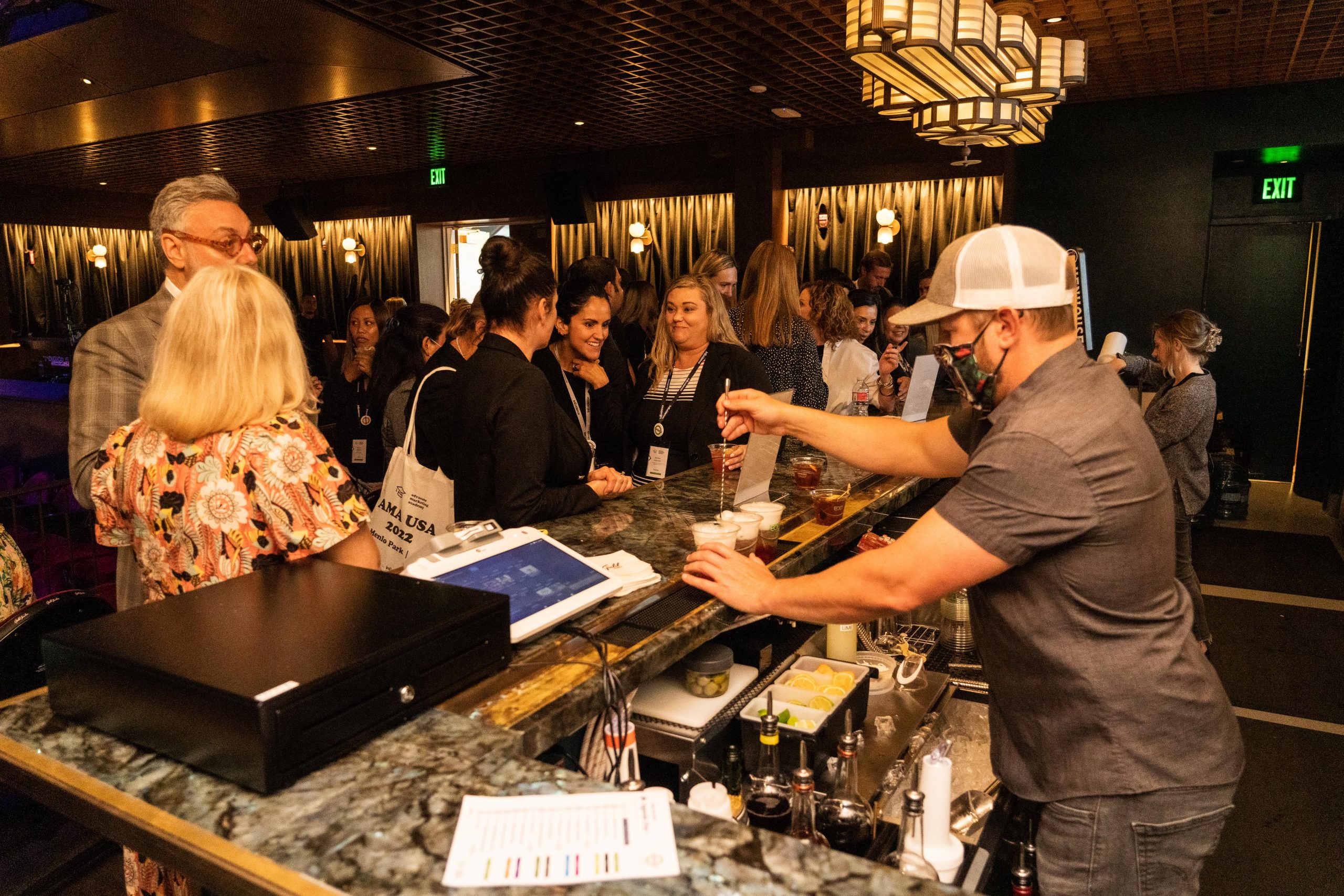 A busy bar scene with a bartender preparing a drink for patrons gathered around the bar counter. The bar has a marble countertop and various drink ingredients. Many people are engaged in conversation, and the ambiance is lively with warm lighting and contemporary decor.