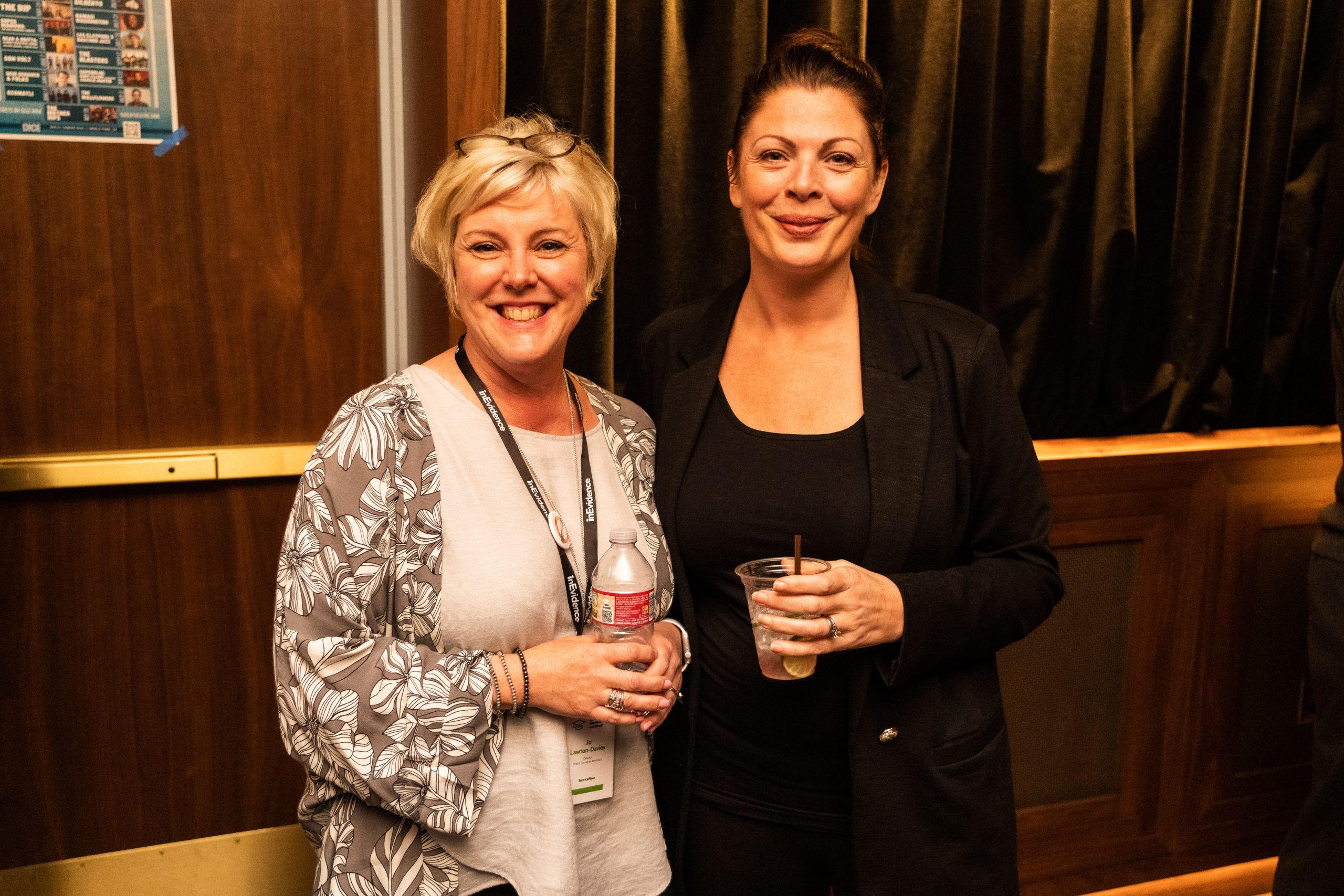 Two women standing together indoors, smiling at the camera. One woman has short blond hair, wears a patterned grey cardigan over a white top, and holds a water bottle. The other woman has dark, pulled-back hair, wears a black outfit, and holds a drink with a straw.