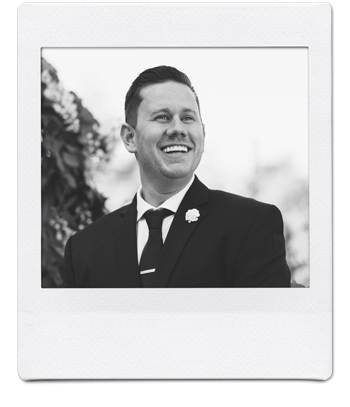 Black and white photo of a smiling man wearing a suit and tie, with a flower on his lapel, framed in a Polaroid-style border. He stands outdoors with foliage in the background.