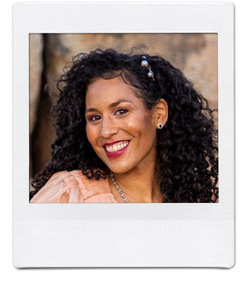 A woman with curly dark hair adorned with beads, wearing a peach-colored top and earrings, smiles brightly at the camera in a photo with a white border. The background is blurred but shows hints of a natural outdoor setting.