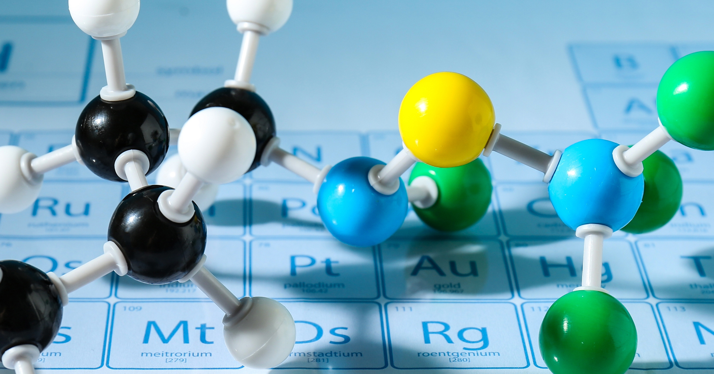 Colorful molecular models, consisting of black, white, green, blue, and yellow spheres connected by rods, are placed on a blue periodic table of elements. The image highlights the intersection of chemistry and molecular structures while subtly showcasing customer advocacy in scientific education.
