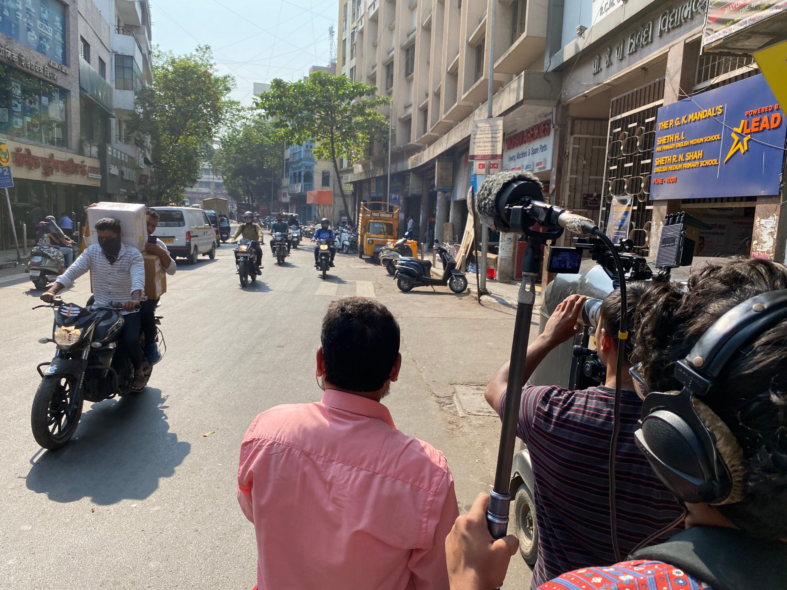 A busy urban street scene with motorcyclists riding in the sunlight. A camera crew films from the side of the road, focusing on a biker wearing a white shirt. Tall buildings line the street, with parked vehicles and shops visible.