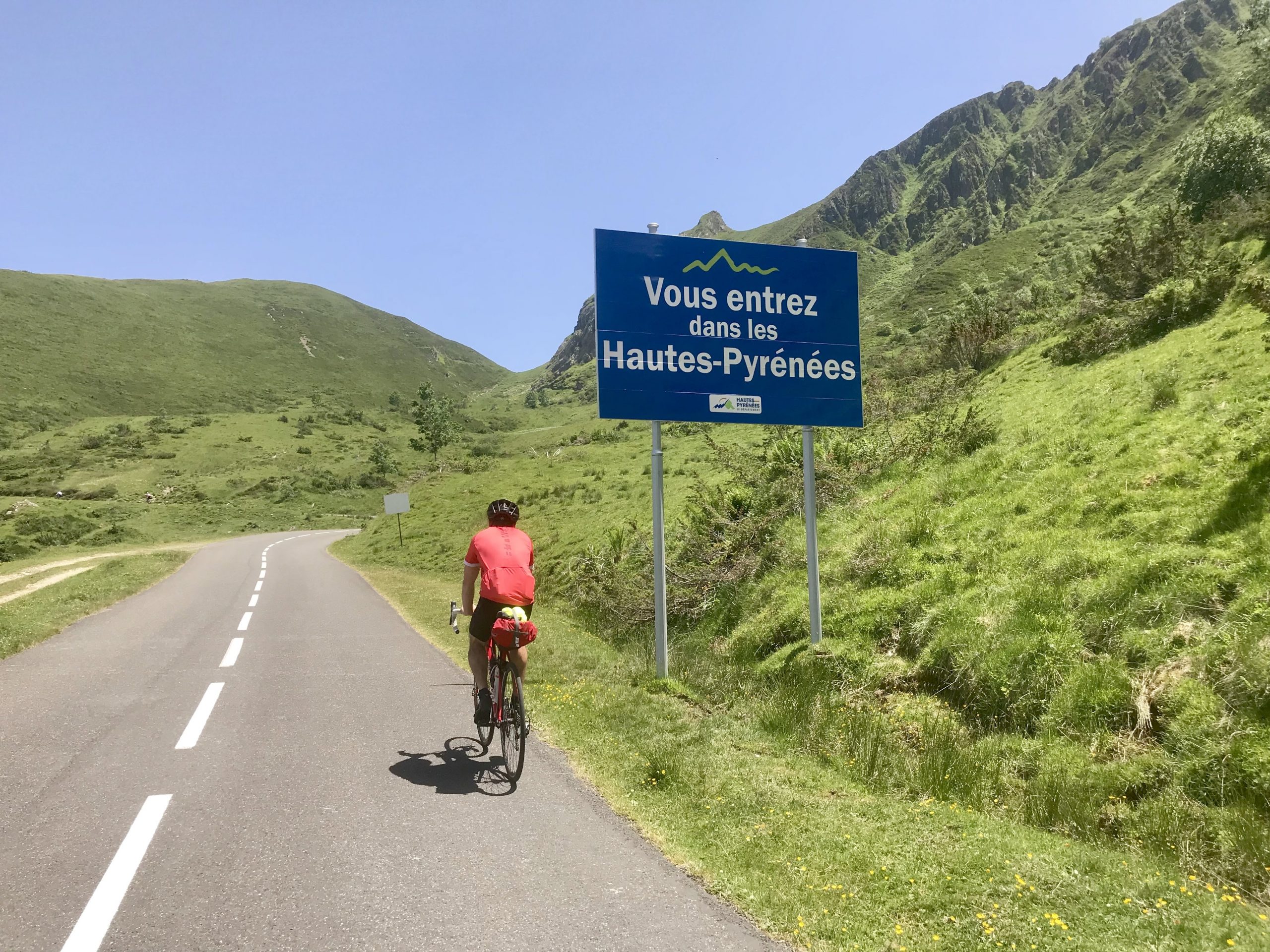 A cyclist in a red outfit rides up a paved road next to a sign that reads "Vous entrez dans les Hautes-Pyrénées" amidst lush green hills and a clear blue sky.