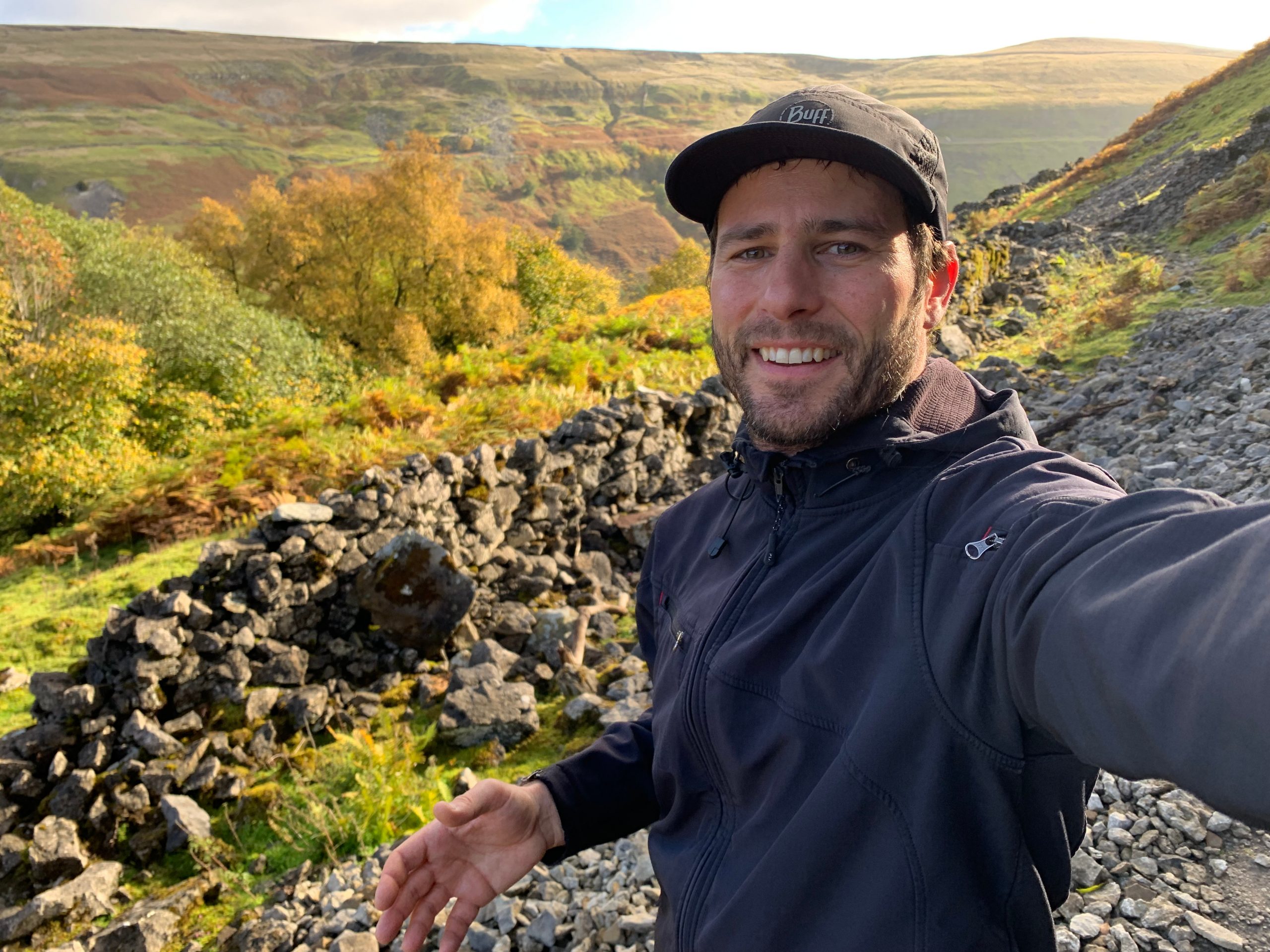 A person takes a selfie while hiking in a scenic, hilly landscape. They wear a black jacket and cap, smiling with rocky terrain and green, autumn-colored foliage in the background.