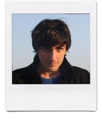 A person with dark, tousled hair and wearing a dark jacket over a blue top is staring directly at the camera with a neutral expression. The background is a clear sky. The image is framed to resemble a polaroid photo.