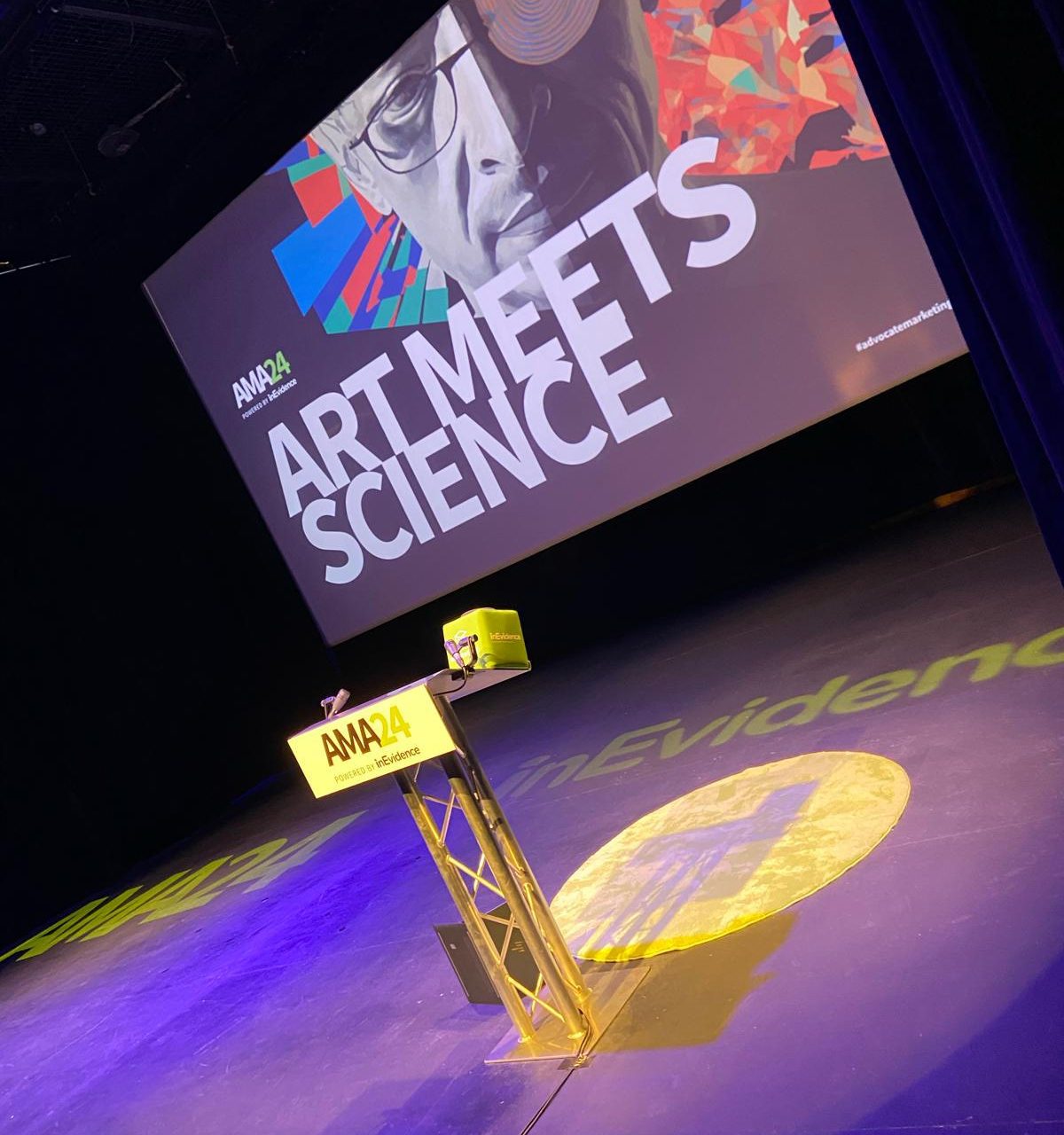 A stage with a podium and a large screen displaying "AMA24 ART MEETS SCIENCE" along with a colorful image of a face. The podium has a small yellow box on it and a projected light circle on the floor with text partially visible.